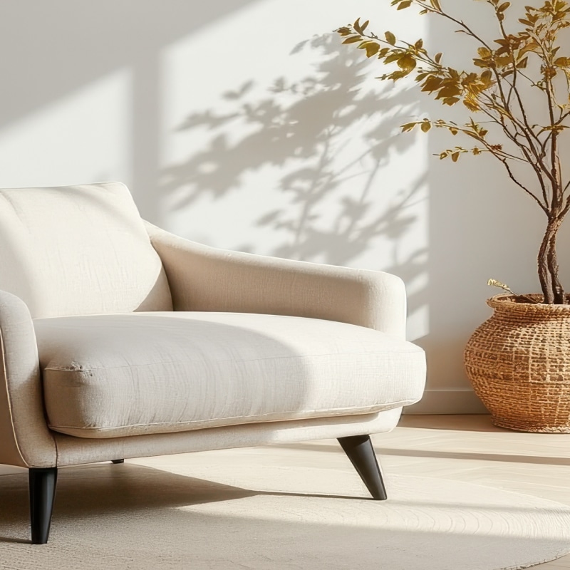 Fauteuil Beige Confortable : Lumière et Ombre Naturelles Fauteuil beige clair moderne dans un intérieur lumineux, projetant des ombres d'arbres sur le mur blanc à côté d'une plante en panier tressé.