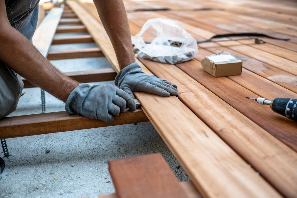 Installation de terrasse en bois par un professionnel Ouvrier ganté posant des lames de platelage en bois exotique sur une terrasse. Les outils (perceuse et vis) sont prêts pour la construction.